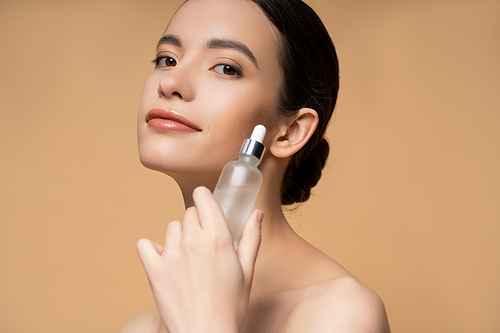 Pleased young asian woman with naked shoulders holding cosmetic serum isolated on beige