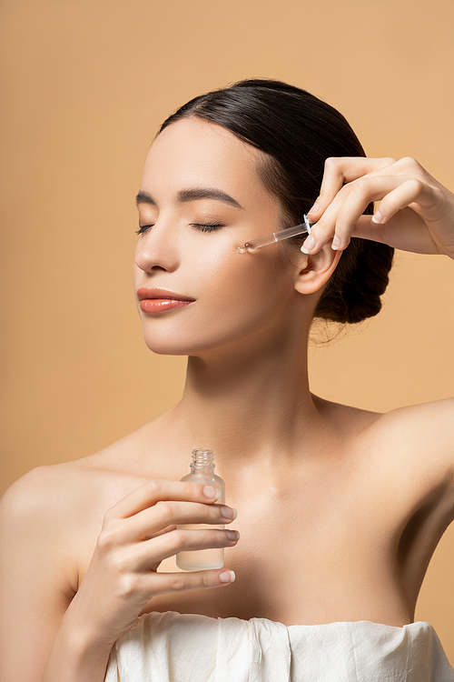 Pretty asian woman with closed eyes in top applying cosmetic serum while posing isolated on beige
