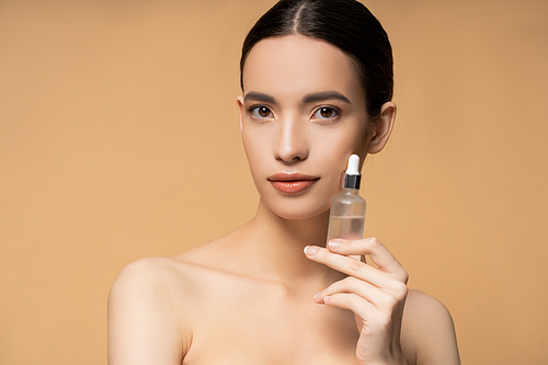 Pretty asian woman with naked shoulders holding cosmetic serum and standing isolated on beige