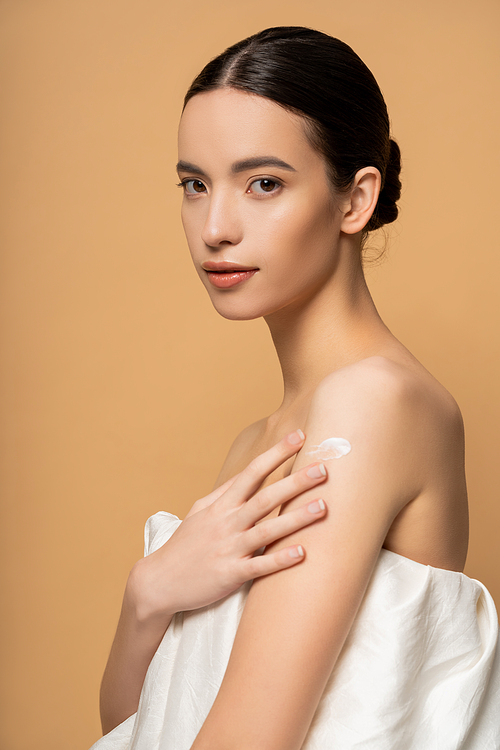 Young brunette asian woman applying body cream on shoulder while standing isolated on beige