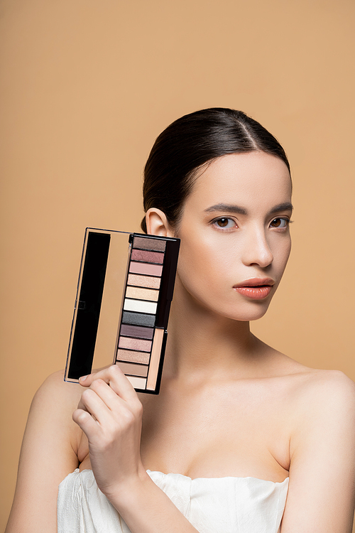 Pretty young asian woman in top with naked shoulders holding eyeshadow palette isolated on beige