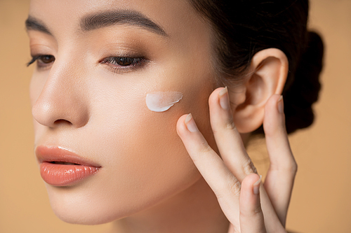 Close up view of young asian woman applying face cream on cheek isolated on beige
