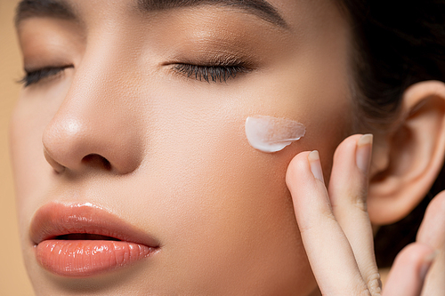 Close up view of young asian woman with perfect skin applying face cream on cheek isolated on beige