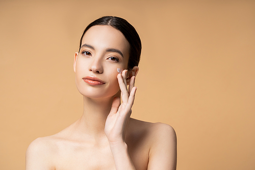 Young asian woman looking at camera while applying face cream and standing isolated on beige