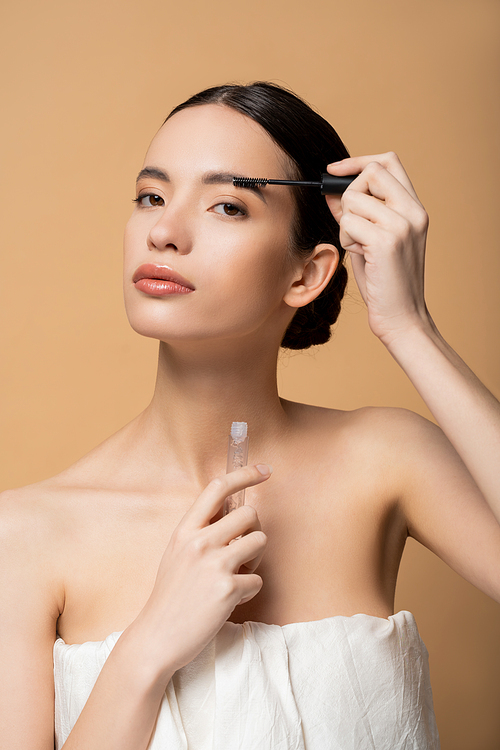 Confident young asian woman in top applying eyebrow gel and looking at camera isolated on beige