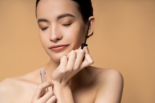 Young asian woman with naked shoulders and closed eyes holding mascara isolated on beige