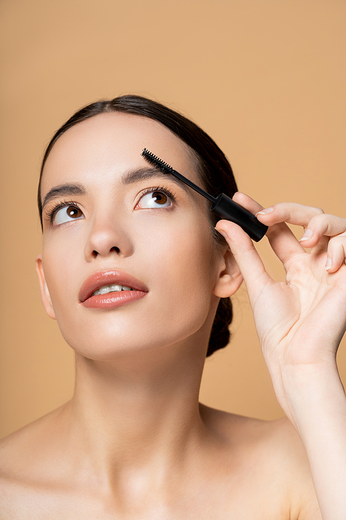 Pretty young asian woman with naked shoulders looking up and apply eyebrow gel isolated on beige