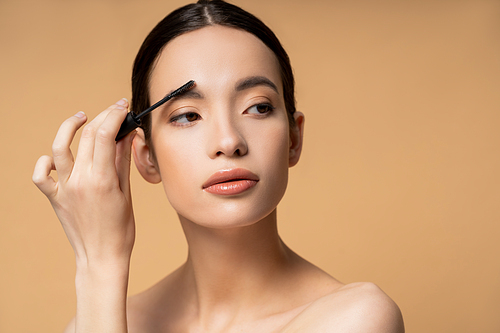 Young brunette asian woman with naked shoulders applying eyebrow gel while posing isolated on beige