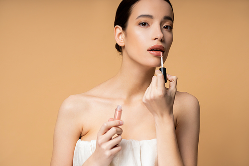 Pretty young asian woman in top looking at camera while applying lip gloss isolated on beige