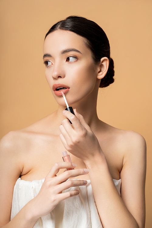 Pretty asian model with naked shoulders in top applying lip gloss and looking away isolated on beige
