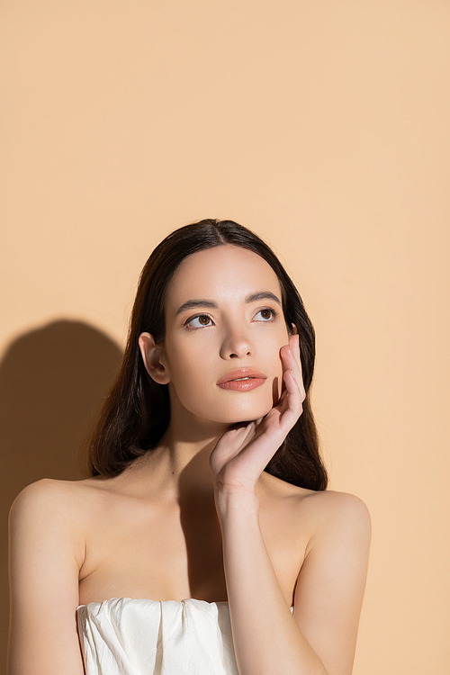 Asian woman with naked shoulders touching cheek while standing on beige background with shadow