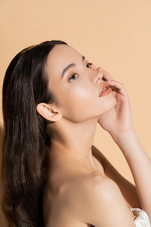 Pretty asian woman with natural makeup touching cheek while standing on beige background