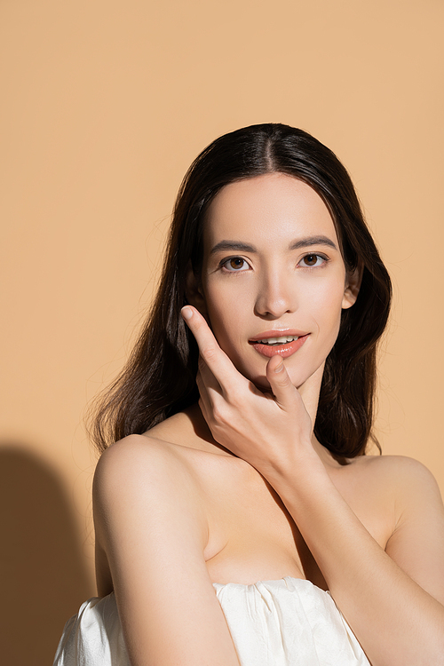 Smiling brunette asian woman in top touching cheek and looking at camera on beige background