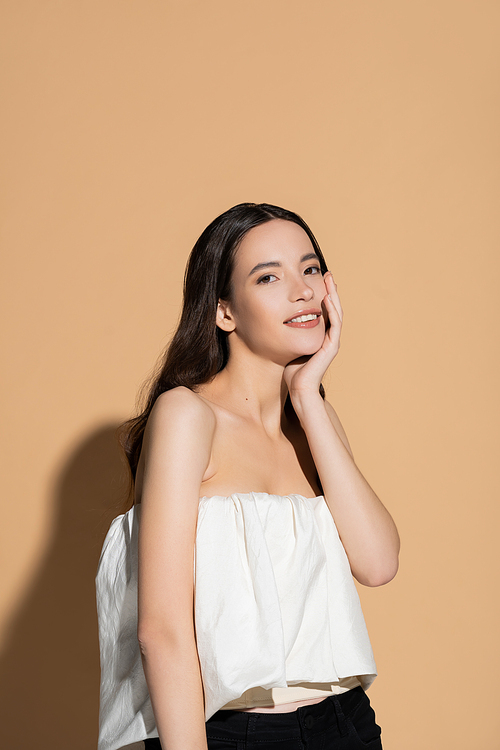 Beautiful asian woman in top touching face and smiling at camera on beige background with shadow