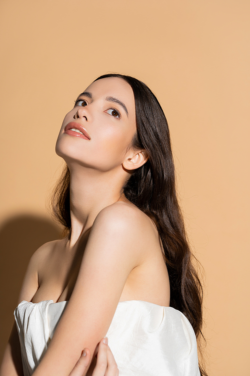 Dreamy young asian woman in top with naked shoulders standing on beige background with shadow