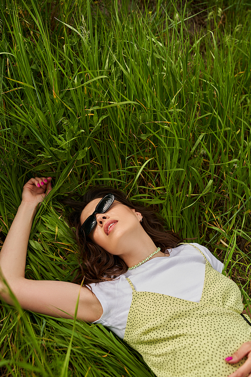 Top view of fashionable brunette woman in sunglasses and sundress lying on green grass on field at summer, natural landscape  and relaxing in nature concept, rural landscape