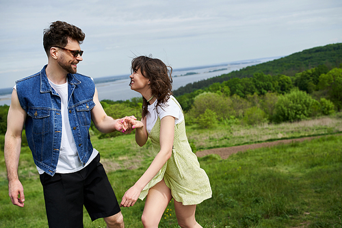 Smiling and stylish man in sunglasses holding hand of cheerful girlfriend in sundress while spending time and standing together with scenic landscape at background, couple in love enjoying nature