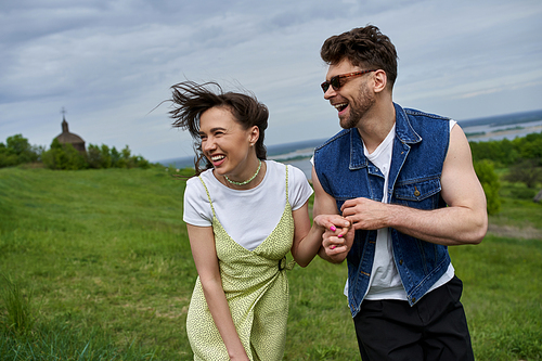 Cheerful bearded man in sunglasses and denim vest holding hand of brunette girlfriend in sundress while walking on grassy field, couple in love enjoying nature and relaxing concept