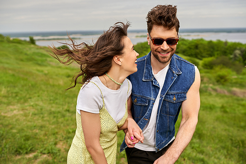 Cheerful brunette woman in sundress holding hand of stylish boyfriend in sunglasses and denim vest and walking on blurred grassy field, couple in love enjoying nature, tranquility