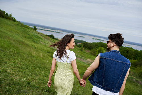 Side view of smiling and trendy romantic couple in summer outfits holding hands while walking on grassy hills with cloudy sky at background, couple in love enjoying nature, tranquility