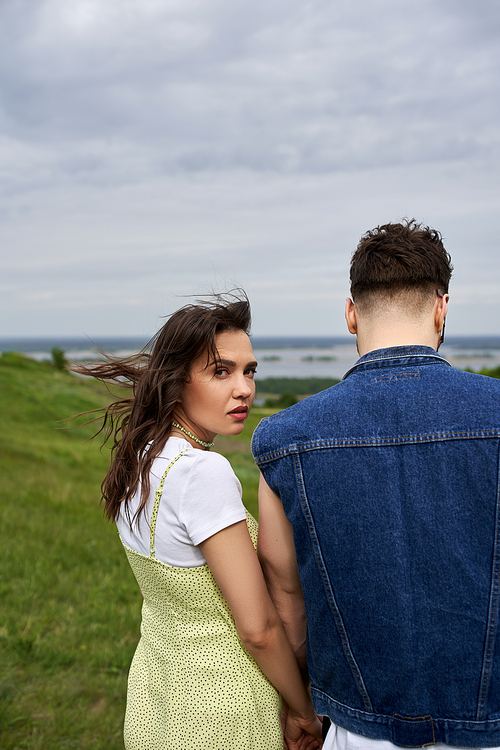 Fashionable brunette woman in summer sundress looking at camera and holding hand of boyfriend in denim vest and standing on blurred green field, couple in love enjoying nature, tranquility