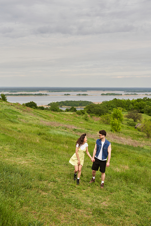 Cheerful brunette woman in sundress and boots talking to stylish boyfriend in sunglasses and denim vest and walking on hill with grass together, couple in love enjoying nature, tranquility