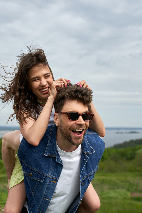 Cheerful brunette woman piggybacking on stylish boyfriend in sunglasses and denim vest while standing with blurred landscape at background, countryside adventure and love story, tranquility