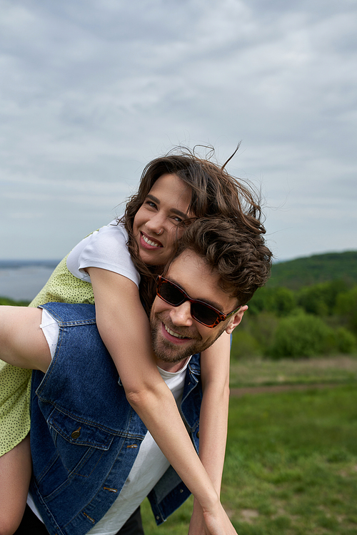 Cheerful brunette woman in sundress hugging stylish boyfriend in sunglasses and denim vest while piggybacking and looking at camera with landscape at background, countryside adventure and love story
