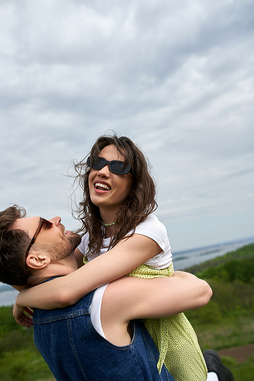 Cheerful bearded man in denim vest ad sunglasses hugging stylish brunette girlfriend in sundress and spending time in rural landscape at background, love story and countryside adventure