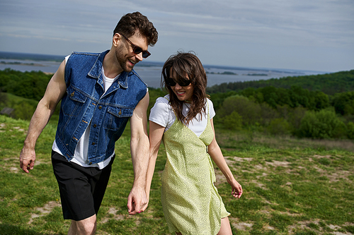 Positive love couple in sunglasses and stylish summer outfits holding hands while spending time on blurred landscape at background, countryside wanderlust and love concept, tranquility