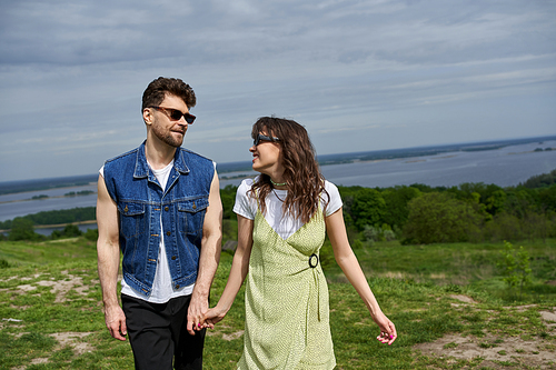 Cheerful brunette woman in sunglasses and stylish sundress holding hand and looking at boyfriend in denim vest and standing on grassy hill, countryside wanderlust and love concept, tranquility