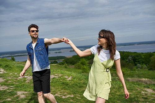 Smiling brunette man in sunglasses and denim vest holding hand of stylish girlfriend in sundress and spending time on grassy hill with cloudy sky at background, countryside wanderlust and love concept