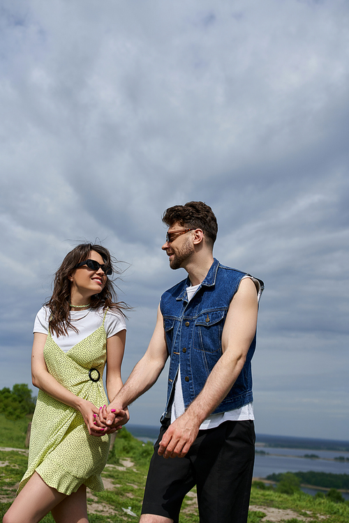 Smiling brunette woman in sunglasses and stylish sundress spending time with bearded boyfriend in summer outfit and denim vest and walking in rural setting, countryside wanderlust and love concept