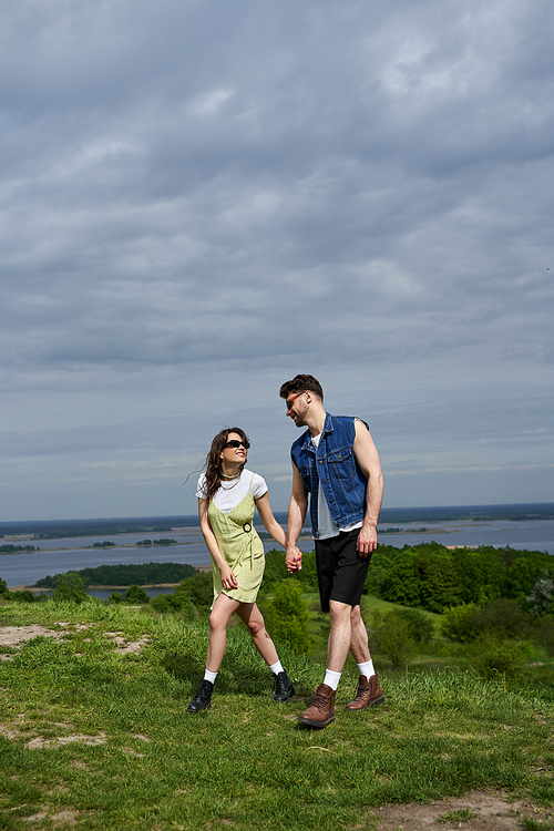 Positive brunette romantic couple in sunglasses and stylish summer outfits holding hands and walking together on grassy hill with blurred scenic view at background, countryside leisurely stroll