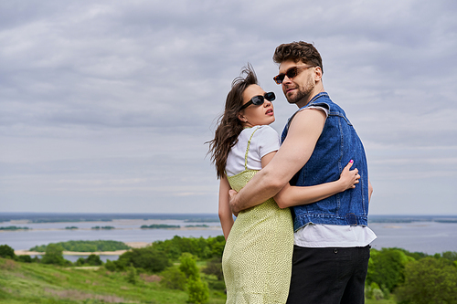 Fashionable romantic couple in sunglasses and summer outfits embracing and standing with blurred rural landscape and cloudy sky at background, countryside leisurely stroll, tranquility