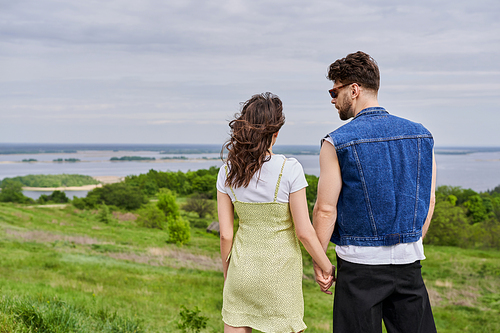 Stylish bearded man in sunglasses and denim vest holding hand of brunette girlfriend in sundress and spending time together on blurred grassy hill at background, countryside retreat concept