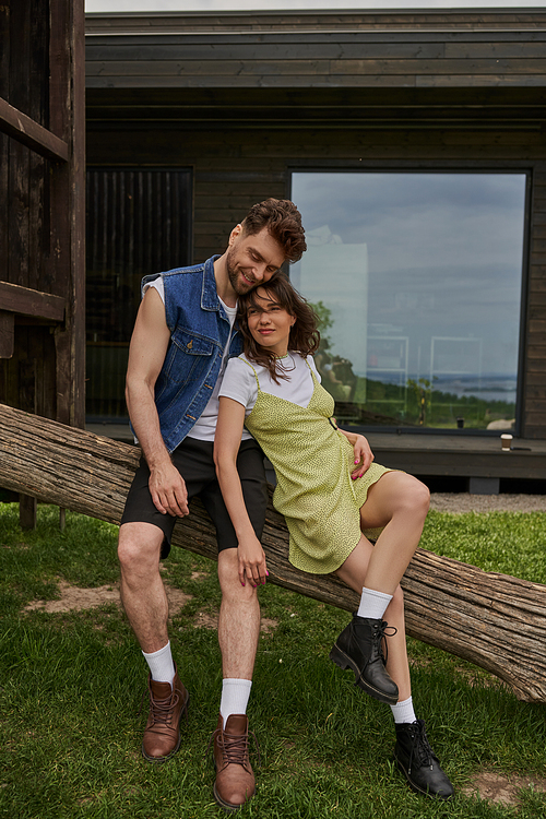 Full length of stylish and fashionable romantic couple in summer outfits and boots resting on wooden log with house at background in rural setting, outdoor enjoyment concept, tranquility