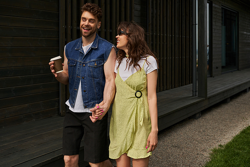 Smiling and stylish bearded man holding coffee to go and hand of brunette girlfriend in sunglasses and summer dress while walking near house in rural setting, outdoor enjoyment concept, tranquility