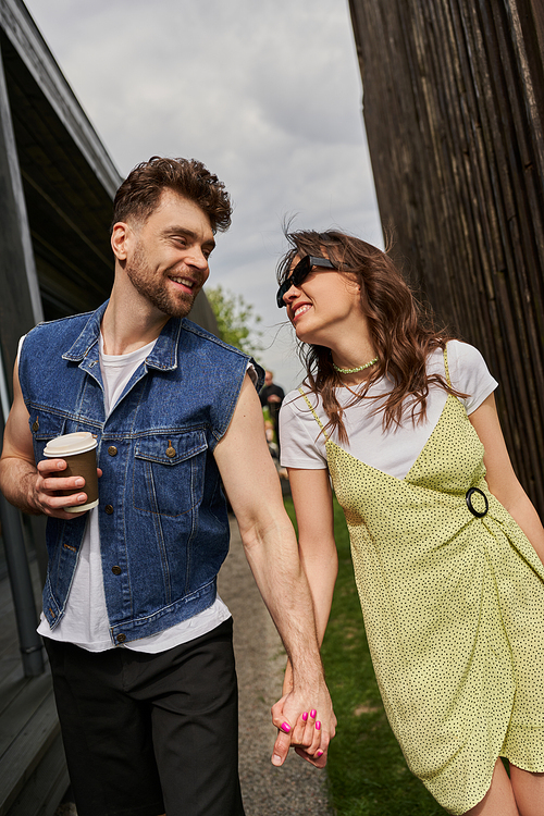Cheerful romantic couple in stylish summer outfits holding hands and coffee to go while talking and walking between wooden houses in rural setting at background, outdoor enjoyment concept