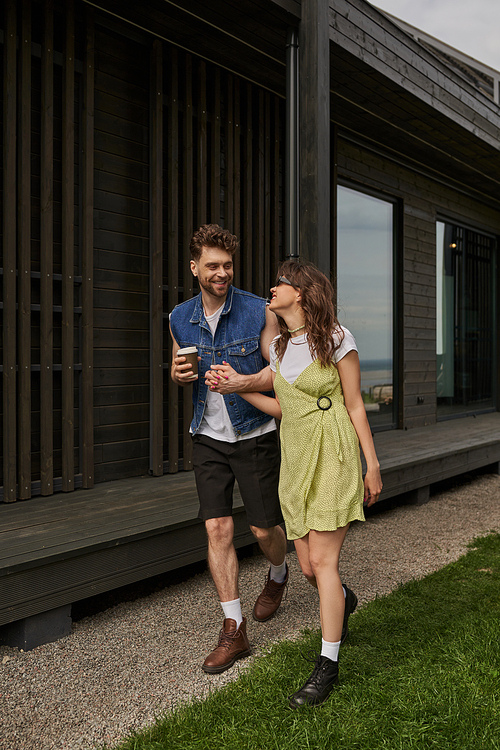 Trendy and stylish couple in summer outfits and boots holding takeaway coffee and talking while walking together near wooden house in rural setting, outdoor enjoyment concept