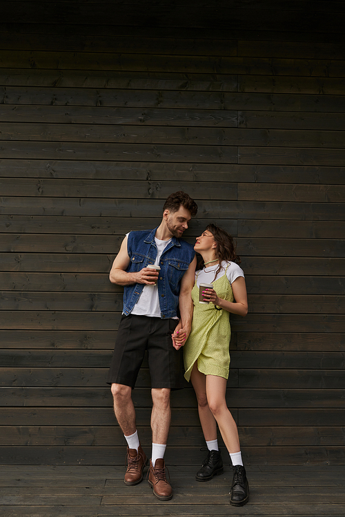 Full length of fashionable romantic couple in boots and summer outfits holding coffee to go and looking at each other while standing near wooden house, carefree moments concept