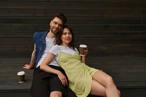 Fashionable stylish and romantic couple in summer outfits smiling at camera while holding coffee to go and sitting near wooden house in rural setting, carefree moments concept, tranquility
