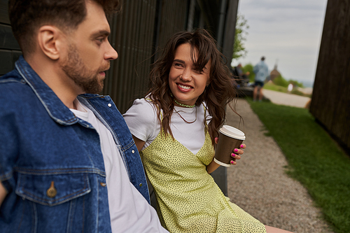 Positive brunette woman in stylish summer outfit holding coffee to go and looking at blurred bearded boyfriend in denim vest near wooden house in rural setting, carefree moments concept