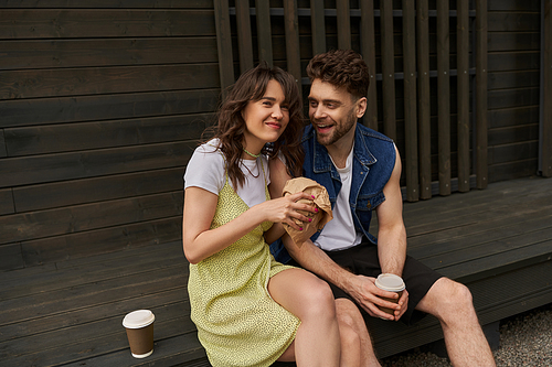 Cheerful brunette woman in sundress holding craft package and looking at camera near bearded boyfriend with coffee to go and wooden house in rural setting, serene ambiance concept