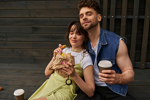 Positive bearded man in denim vest holding coffee to go and sitting near stylish brunette girlfriend in sundress holding tasty bun and wooden house at background, serene ambiance concept