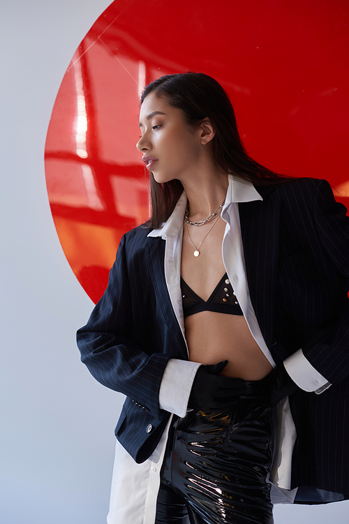 edgy fashion, young asian woman in bra, white shirt and blazer posing in latex pants and black gloves and latex pants near red round shaped glass, grey background, underwear and jacket