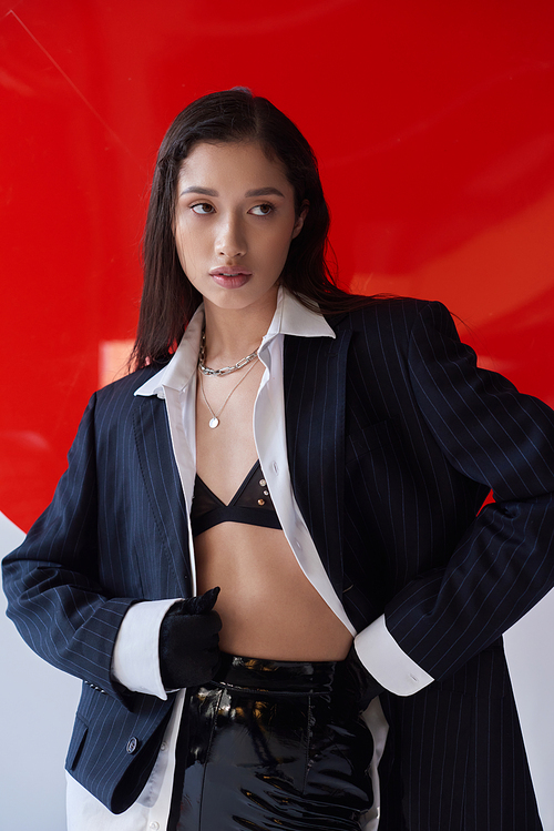 stylish look, young asian woman in bra, white shirt and blazer posing in gloves near red round shaped glass, looking away on grey background, personal style, underwear and jacket, youth