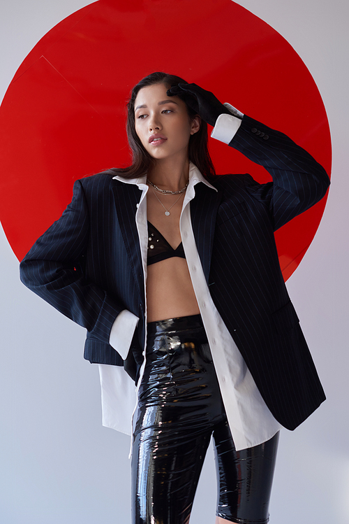 stylish outfit, young asian woman in bra, white shirt and blazer posing in gloves and latex shorts near red round shaped glass, looking away on grey background, personal style, youth