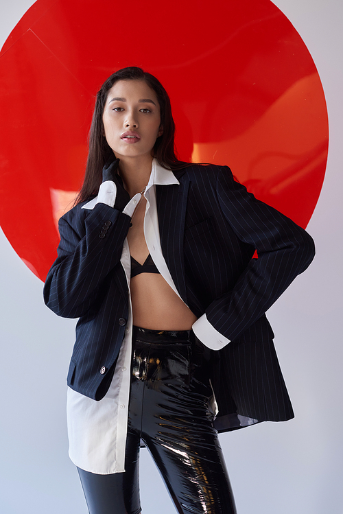 fashion photography, young asian model in bra, white shirt and blazer posing in gloves and latex shorts near red round shaped glass, grey background, looking away, personal style, youth trend