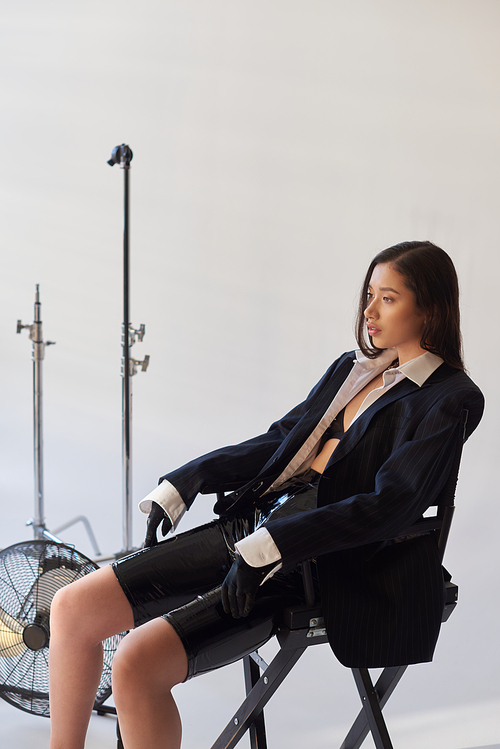 studio photography, young asian model in blazer, white shirt and latex shorts sitting on folding chair near electric fan on grey background, fashion and style, looking away, full length